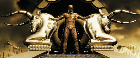 Xerxes stands, arms spread, atop his throne, featuring two antelopes of gold. He is covered in gold piercings and adornments.