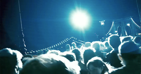 The heads of people moving across a bridge leading out of the city, lit by a helicopter light.