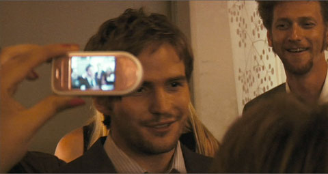 Rob smiling in front of friends. One of them is behind him smiling, and in the foreground is a camera phone held up to capture his speech.