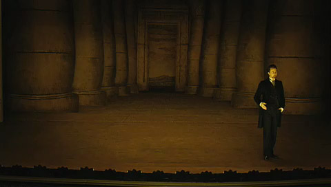 Eisenheim stands on an empty stage in front of a backdrop with fat pillars painted on it in forced perspective.