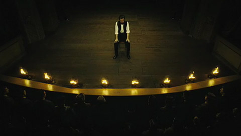 Eisenheim sitting on an older stage, as seen from above, with his sleeves up and his hands on his knees in front of gas footlights.