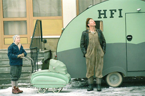 A man and a boy with a baby carriage look to the sky in front of a small camper that reads 'HFI'.