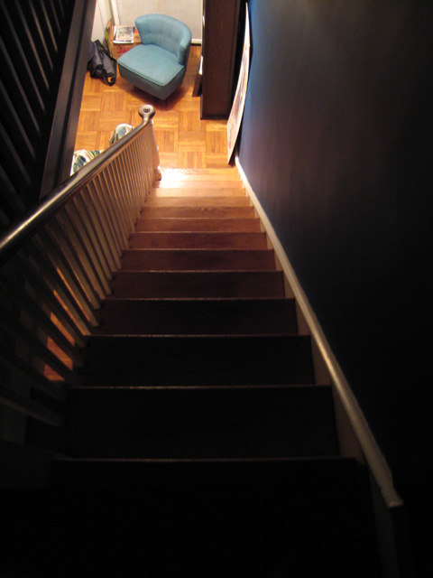 A view down our wooden stairs, which are lit at their foot and get darker as they ascend. On the landing is a bookcase and a turquoise chair.