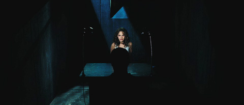 Evey, with a full head of hair, is questioned in a dark room at a table, by a man whose silhouette rises up in front of her.