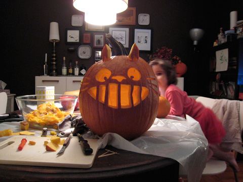 A jack-o-lantern of Totoro's face, sitting on a table covered with implements for carving. Blackbird leans on the table behind the pumpkin.