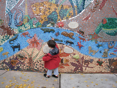 Blackbird, wearing a red coat, walks along a mosaic wall of animals in the jungle, and touches it with her right hand.
