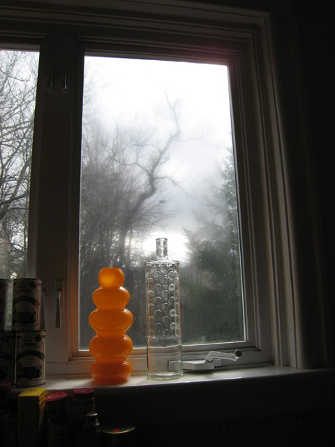 The sunlight hits a bottle and orange vase in the window, which looks out on a hazy, leafless, wooded area.
