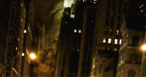 A sliver of a massive shape, covered up by dust a debris, moving behind some buildings at night.