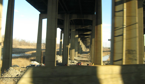 A view underneath a highway overpass, looking straight down the rows of concrete columns that hold the roadway up over the marshes.