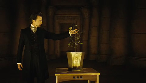 Eisenheim stands with his arm stretched out behind a small tree growing out of a pot on a table.