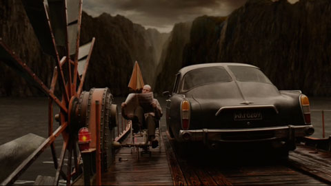 A car is ferried across a bay on a float that is powered by a seated man pedaling large rotors.