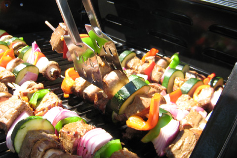Skewers of steak, peppers, mushrooms, red onions, and zucchini on the barbecue, being turned with metal tongs.