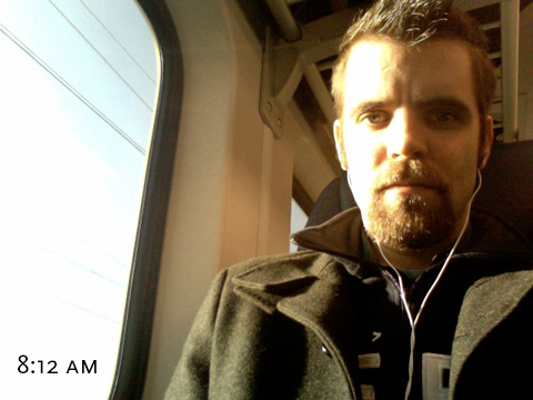 An image of face, light from my right by the rising sun, with a bit of the train window visible. Some power lines can be seen passing by.