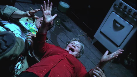 A smiling old woman in a red sweater swings her outstretched hands towards each other as two others reach for them.