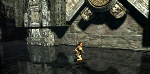 Lara runs in shallow water past a large stone device that looks like a cog.
