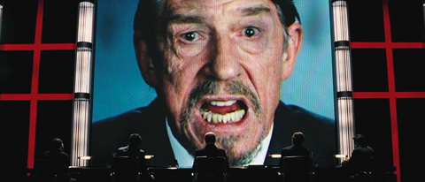 Sutler's giant face looms on a screen over his ministry cabinet members in a dark board room with red Norsefire crosses flanking them.